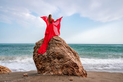 woman sea red dress. Woman with long hair on a sunny seashore in a red flowing dress, back view, silk fabric waving in the wind. Against the backdrop of the blue sky and mountains on the seashore