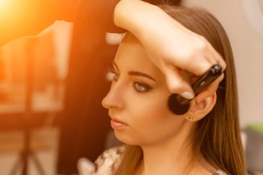 Makeup beauty salon. Makeup artist applies powder and blush. Beautiful female face. Makeup master's hand puts blush on cheeks of beauty model girl. Makeup in progress.