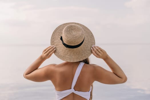 A woman wearing a straw hat is standing on a beach. She is holding her hat up to her face, possibly to shield her eyes from the sun. The beach is calm and peaceful, with the water reflecting the sky