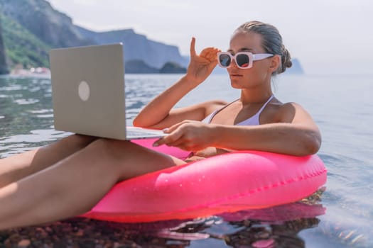 A woman is sitting on a pink inflatable raft in the ocean, using a laptop. Concept of relaxation and leisure, as the woman enjoys her time by the water while working on her laptop