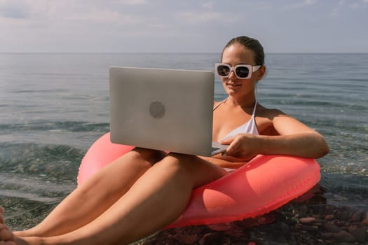 A woman is sitting on a pink inflatable raft with a laptop on her lap. She is wearing sunglasses and a white bikini top. Concept of relaxation and leisure