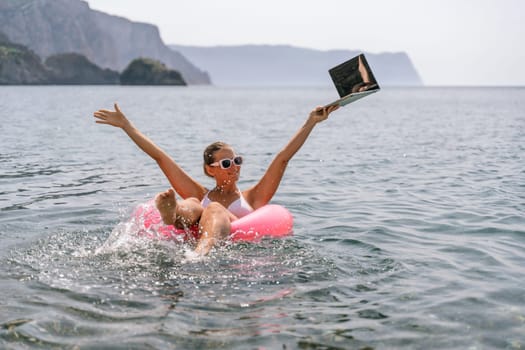 A woman is in a pink inflatable raft in the ocean, holding a laptop. She is smiling and she is enjoying herself