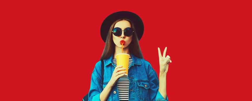 Stylish young woman drinking juice wearing black round hat, jean jacket on red background, blank copy space for advertising text