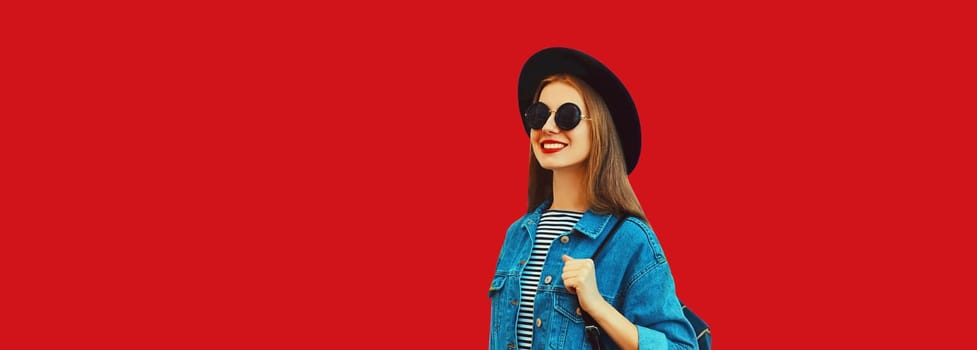 Portrait of happy smiling young woman looking away wearing black round hat, jean jacket, backpack on red background, blank copy space for advertising text