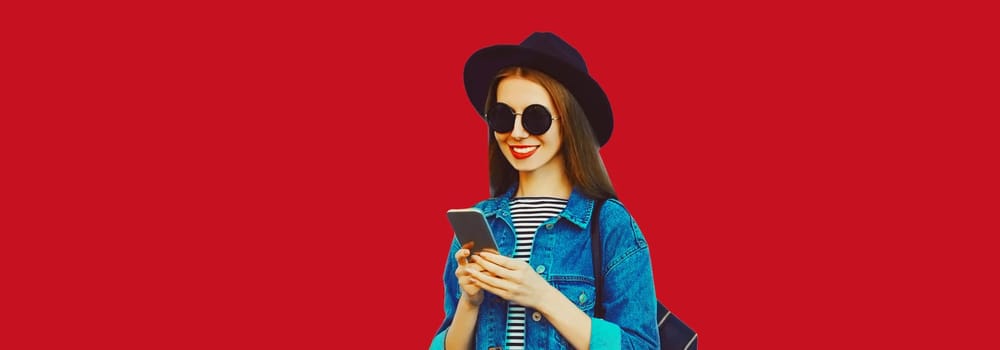 Summer portrait of happy traveler young woman 20s with mobile phone looking at device in black hat with backpack on red studio background