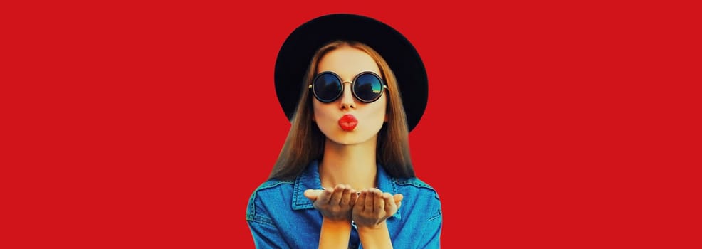 Portrait of stylish young woman model blowing sweet kiss wearing jean jacket, black round hat on red background