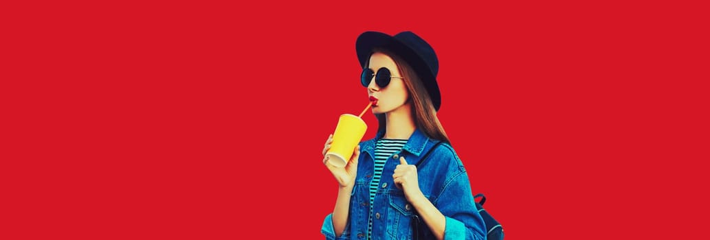 Stylish young woman drinking juice wearing black round hat, jean jacket on red background, blank copy space for advertising text