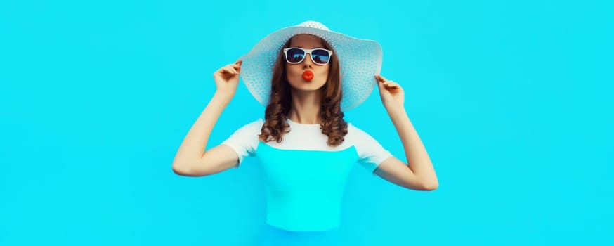 Portrait of beautiful young woman posing blowing kiss wearing summer straw hat and dress on blue background