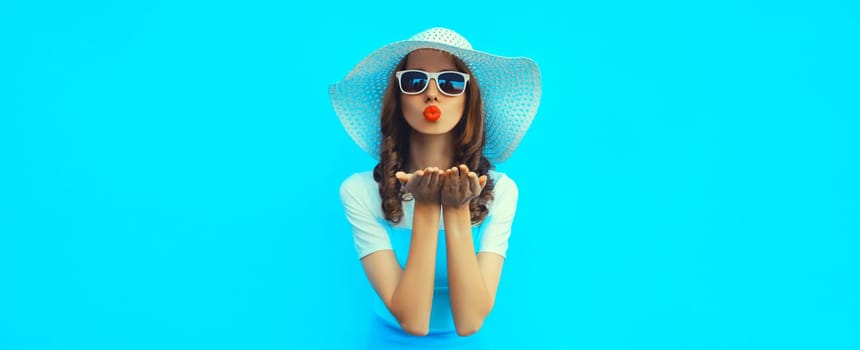 Portrait of beautiful young woman posing blowing kiss wearing summer straw hat and dress on blue background