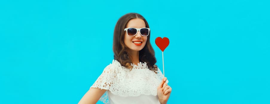 Portrait of beautiful young woman with red heart shaped lollipop on stick wearing white sunglasses on blue studio background