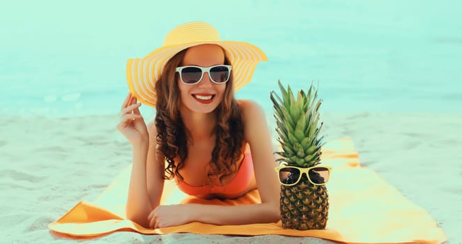 Summer vacation, happy smiling young woman with pineapple fruits in bikini and straw hat lying on sand on the beach on sea background