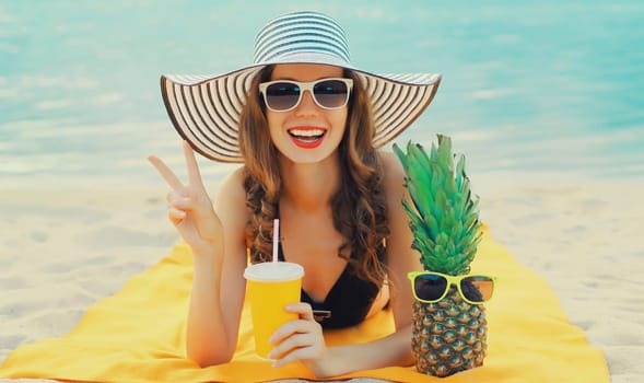 Summer vacation, happy smiling young woman drinks juice with pineapple in straw hat lying on sand on the beach on sea background