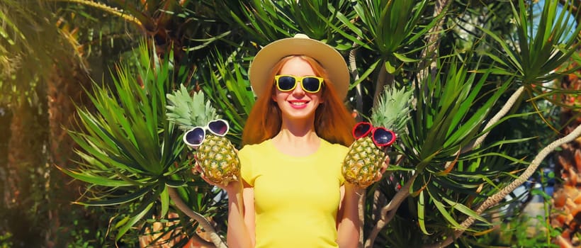 Summer portrait of happy smiling young woman with pineapple fruits wearing sunglasses, straw hat posing on palm tree background