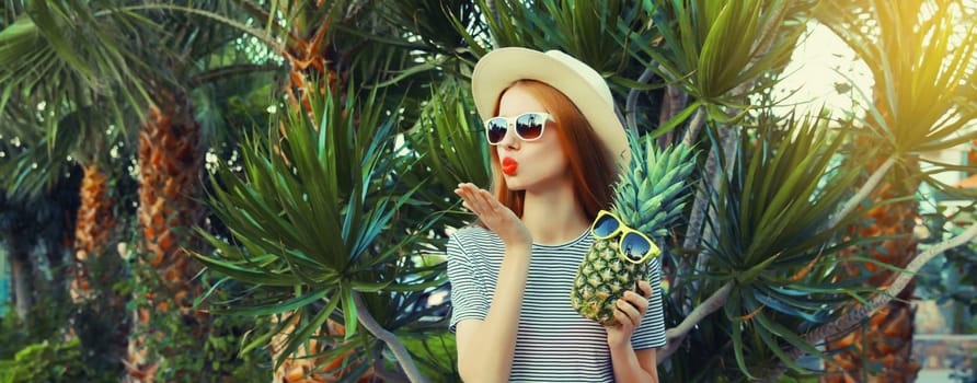 Summer portrait of beautiful young woman with pineapple fruits posing blowing a kiss wearing sunglasses, straw hat on palm tree background