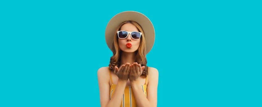 Portrait of beautiful stylish young woman blowing kiss in summer hat, glasses posing on blue background