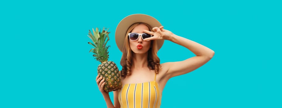 Summer portrait of beautiful young woman blowing a kiss with pineapple wearing straw hat, glasses posing on blue studio background