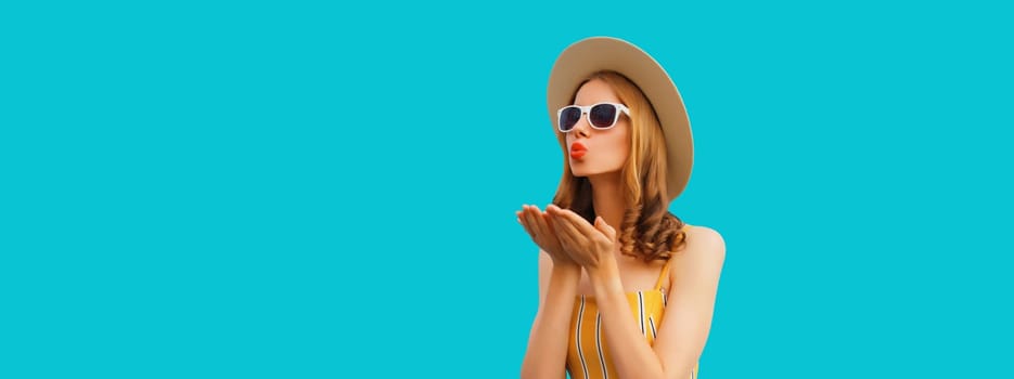 Portrait of beautiful stylish young woman blowing kiss in summer hat, glasses posing on blue background