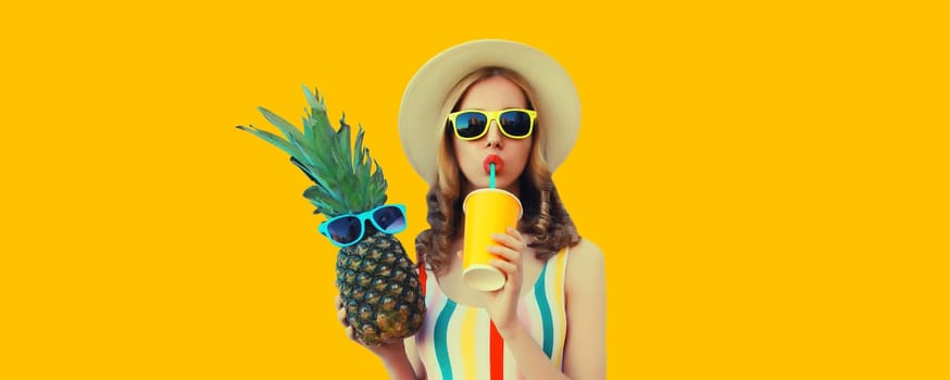 Summer bright portrait of stylish young woman drinking fresh juice holding pineapple fruit in straw hat, sunglasses on colorful yellow studio background