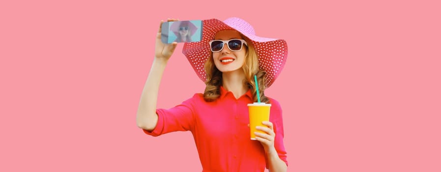 Summer portrait of beautiful happy smiling young woman taking selfie with smartphone and cup of fresh juice wearing straw hat, pink dress on studio background