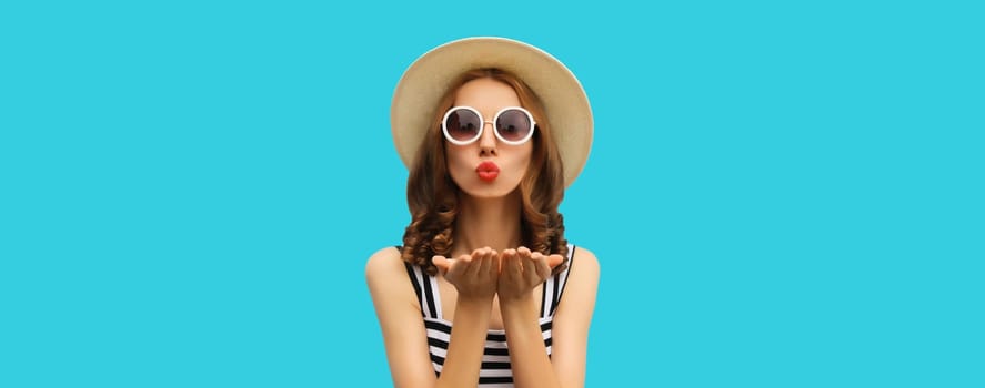 Portrait of beautiful young woman posing blowing kiss wearing summer straw hat and dress on blue background