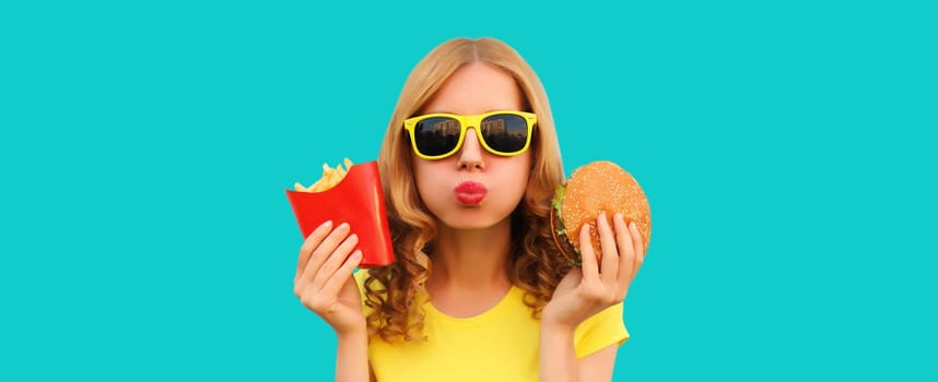 Portrait of happy cheerful young woman eating burger fast food and french fries, fried potatoes isolated on blue studio background