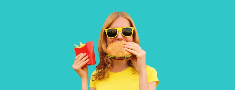 Portrait of happy cheerful young woman eating burger fast food and french fries, fried potatoes isolated on blue studio background