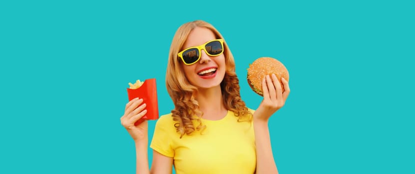 Portrait of happy cheerful young woman eating burger fast food and french fries, fried potatoes isolated on blue studio background