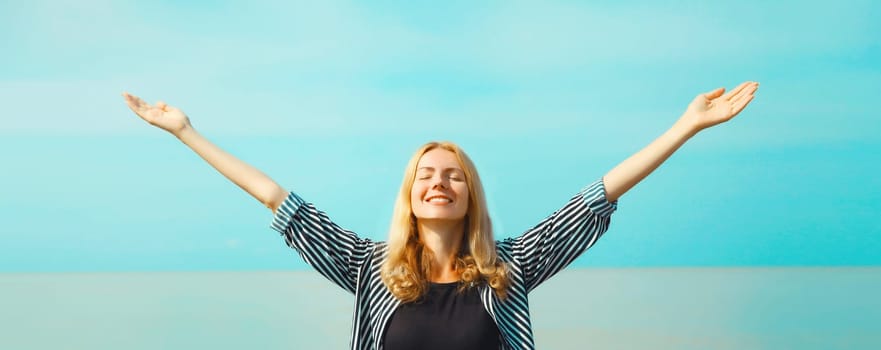 Summer vacation, happy inspired young woman raising her hands up on the beach on sea coast and blue sky background