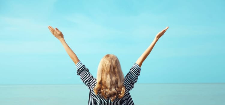 Summer vacation, happy inspired young woman raising her hands up on the beach on sea coast and blue sky background