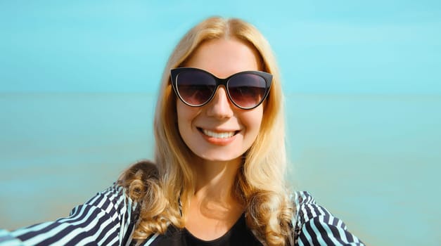 Summer portrait of happy smiling caucasian young woman stretching hand for taking selfie on smartphone on the beach on sea background