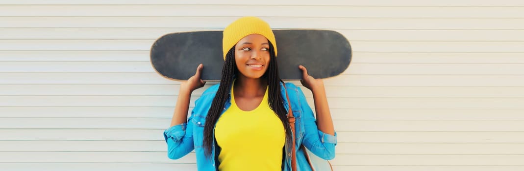 Happy smiling young african woman in colorful yellow hat posing with skateboard in the city on white wall background