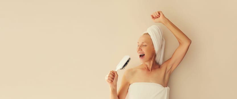 Happy cheerful mature woman having fun sings with hair comb and laughing after shower drying her wet hair with towel wrapped on her head in the morning at home