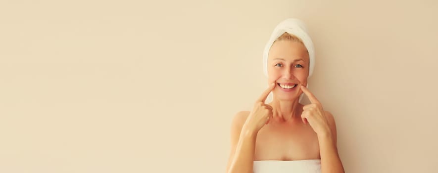 Portrait of happy smiling caucasian middle-aged woman points to her white clean teeth while drying her wet hair with wrapped towel on her head after shower in the morning at home