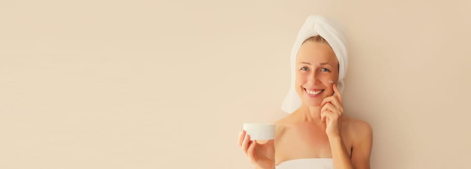 Natural beauty portrait of happy smiling caucasian middle-aged woman touches her clean skin applying face cream drying wet hair with white wrapped bath towel on her head after shower in morning