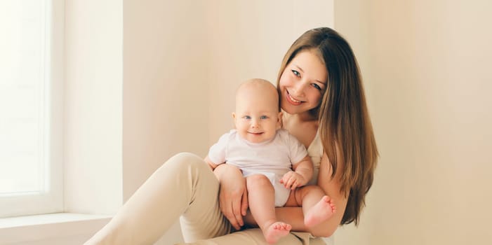 Happy smiling young mother playing with cute baby sitting together in white room at home