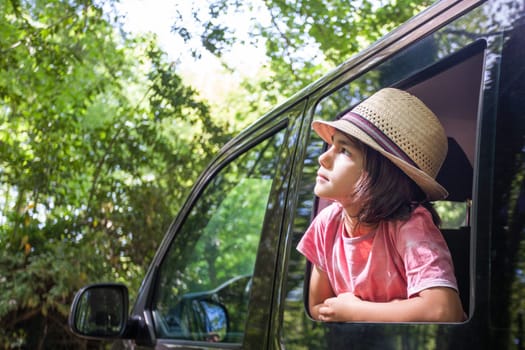 A young boy is sitting in a car with her hat on. He is looking out the window and seems to be enjoying the view