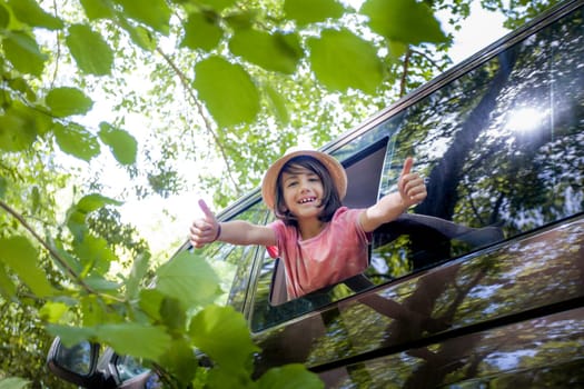 A young boy is giving thumbs up from the window of a car. The car is parked in a forest, and the boy is smiling