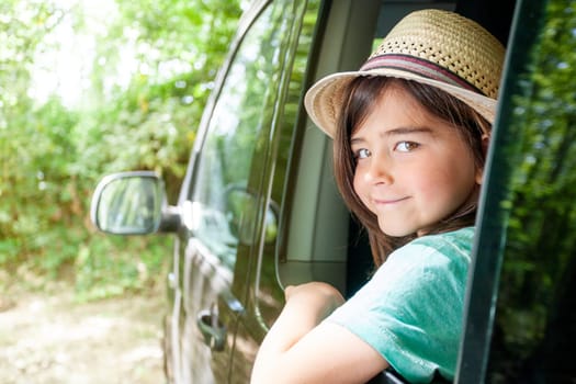 A kid is sitting in a car with her hat on. He is smiling and looking out the window