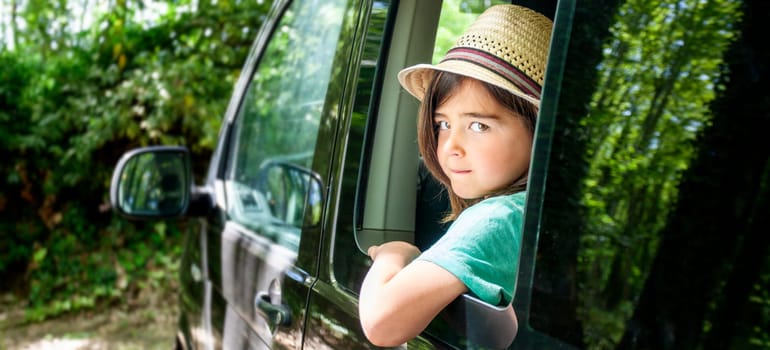 A kid is sitting in a car window, looking out at the trees. Scene is peaceful and calm, as the boy seems to be enjoying the view outside