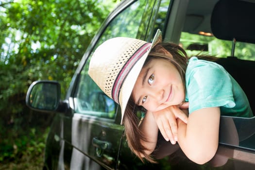 A kid is sitting in a car with her hat on. He is smiling and looking out the window