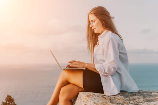 Digital nomad, woman in the hat, a business woman with a laptop sits on the rocks by the sea during sunset, makes a business transaction online from a distance. Freelance, remote work on vacation.