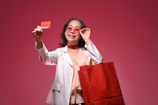Happy smiling senior woman showing credit card holding shopping bags against red background.