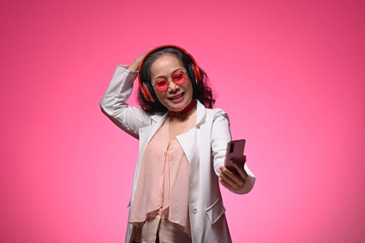 Smiling senior woman holding mobile phone and listening to favourite music over pink background.