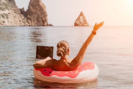 Woman freelancer works on laptop swimming in sea on pink inflatable ring. Pretty lady typing on computer while floating in the sea on inflatable donut at sunset. Freelance, remote work on vacation