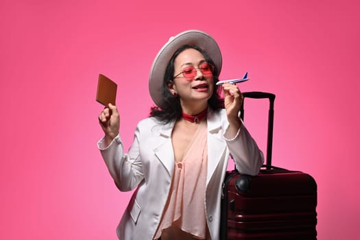 Middle age woman in straw hat holding airplane model and passport on pink background.