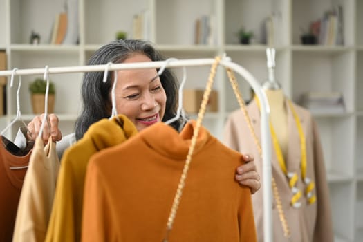 Happy smiling middle female fashionable clothing store owner in apron arranging clothes on rack.