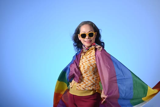 Confident middle woman wearing sunglasses wrapped in a rainbow flag on blue background. LGBT concept.