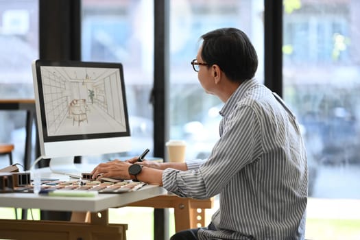 Side view of senior male in eyeglasses working with sketches interior design on computer monitor in office.