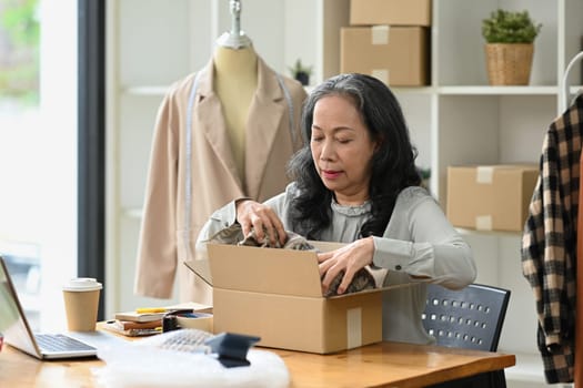 Smiling mature woman ecommerce business worker preparing clothes package at her shop.