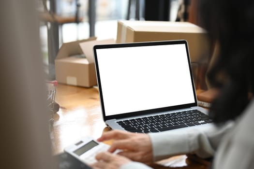 Laptop with blank screen on wooden table with small business owner calculating expenses.